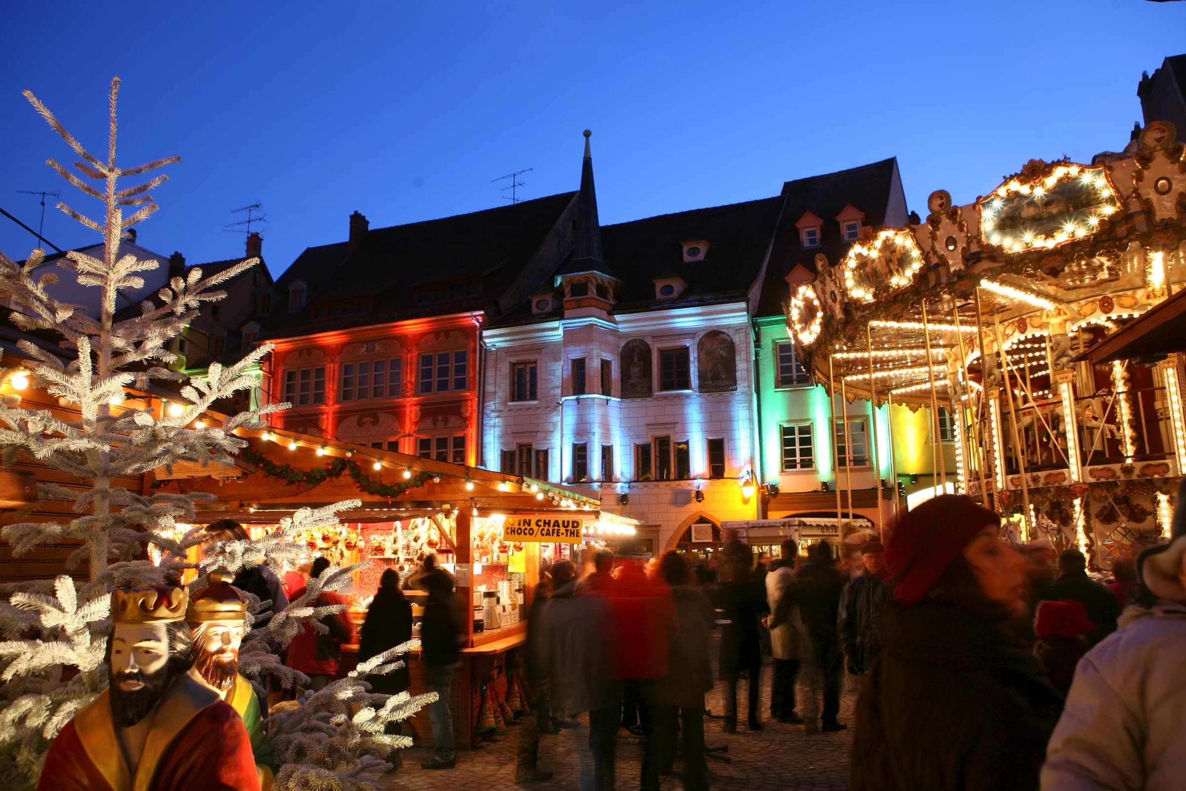 Coloured lights play over the facades of Place de la Réunion in Mulhouse as a carousel turns beside the stalls.