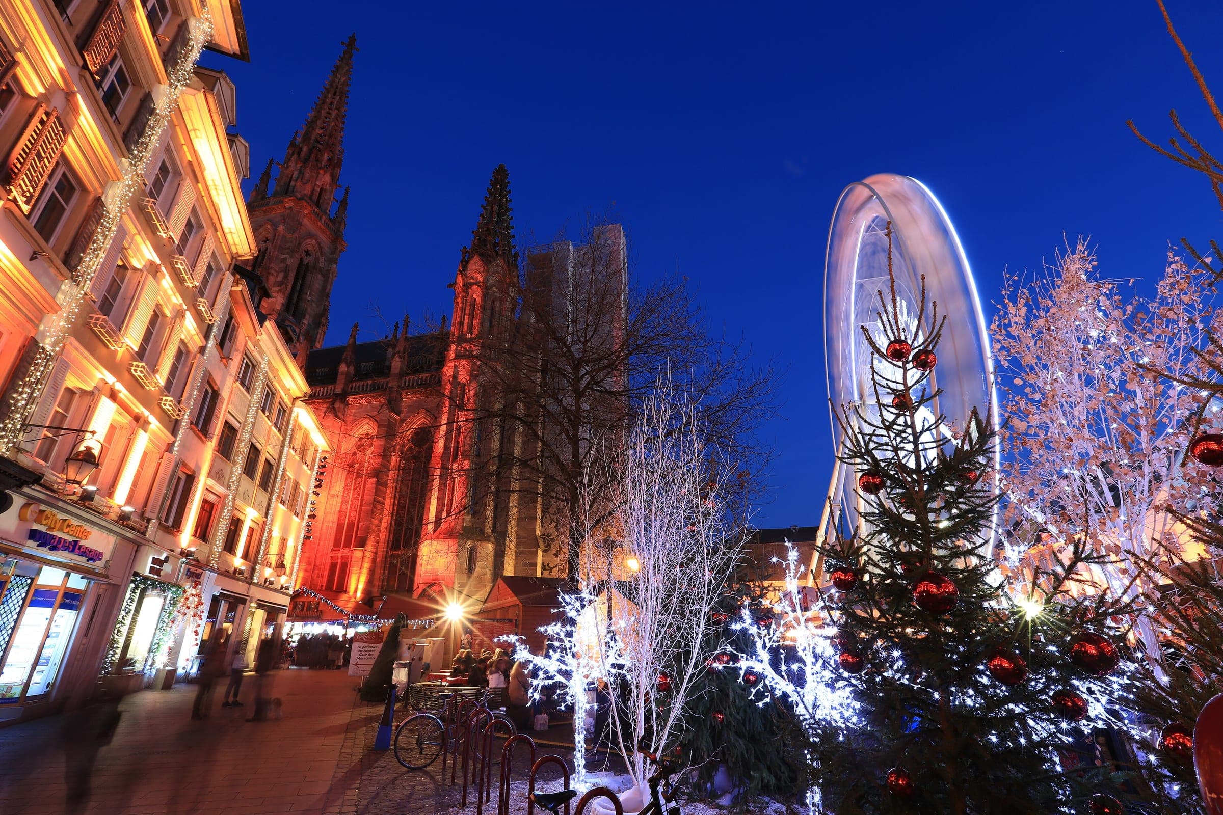 The spire of the Temple Saint-Étienne in Mulhouse rises over lit Christmas trees under a deep blue evening sky.