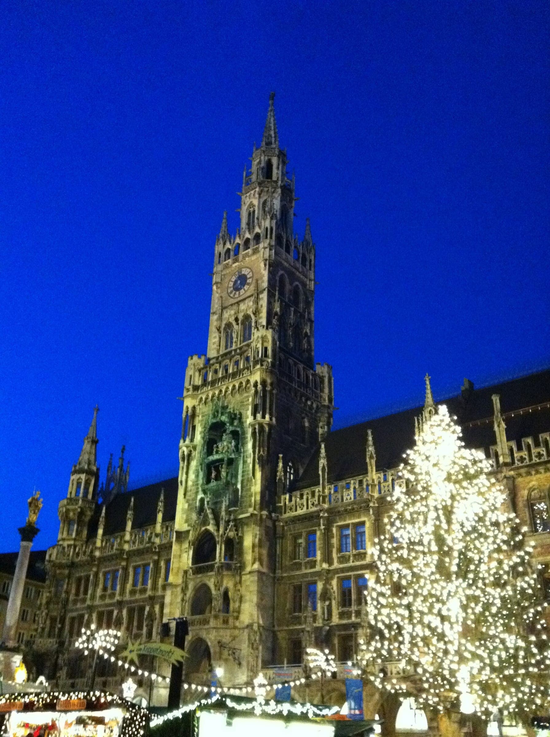 The floodlit tower of Munich's Neues Rathaus rises behind the lit Christmas tree and market huts on Marienplatz.