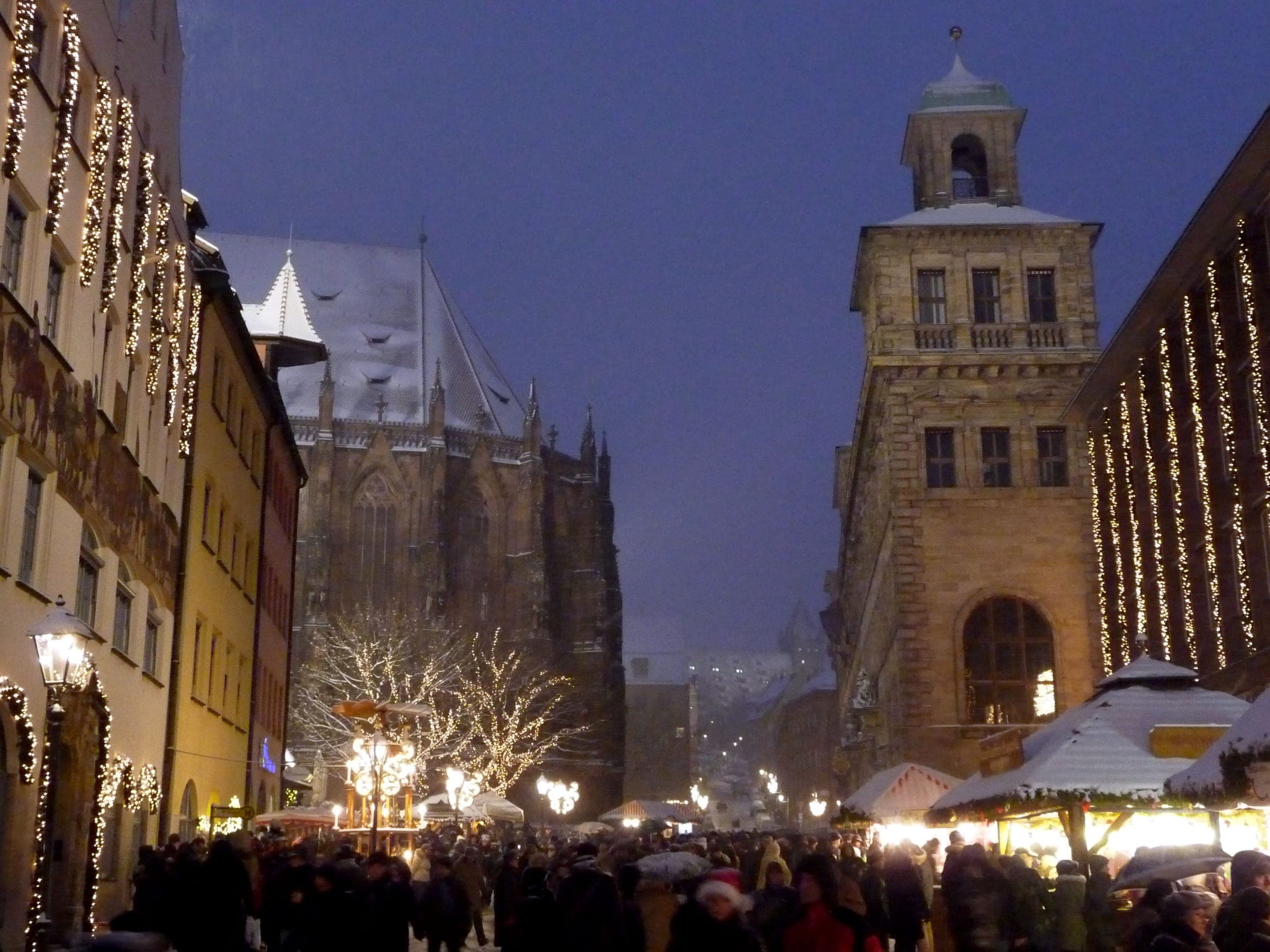 Snow falls on an evening crowd in Nuremberg's old town, with St. Sebald's choir and a lit tower above the street