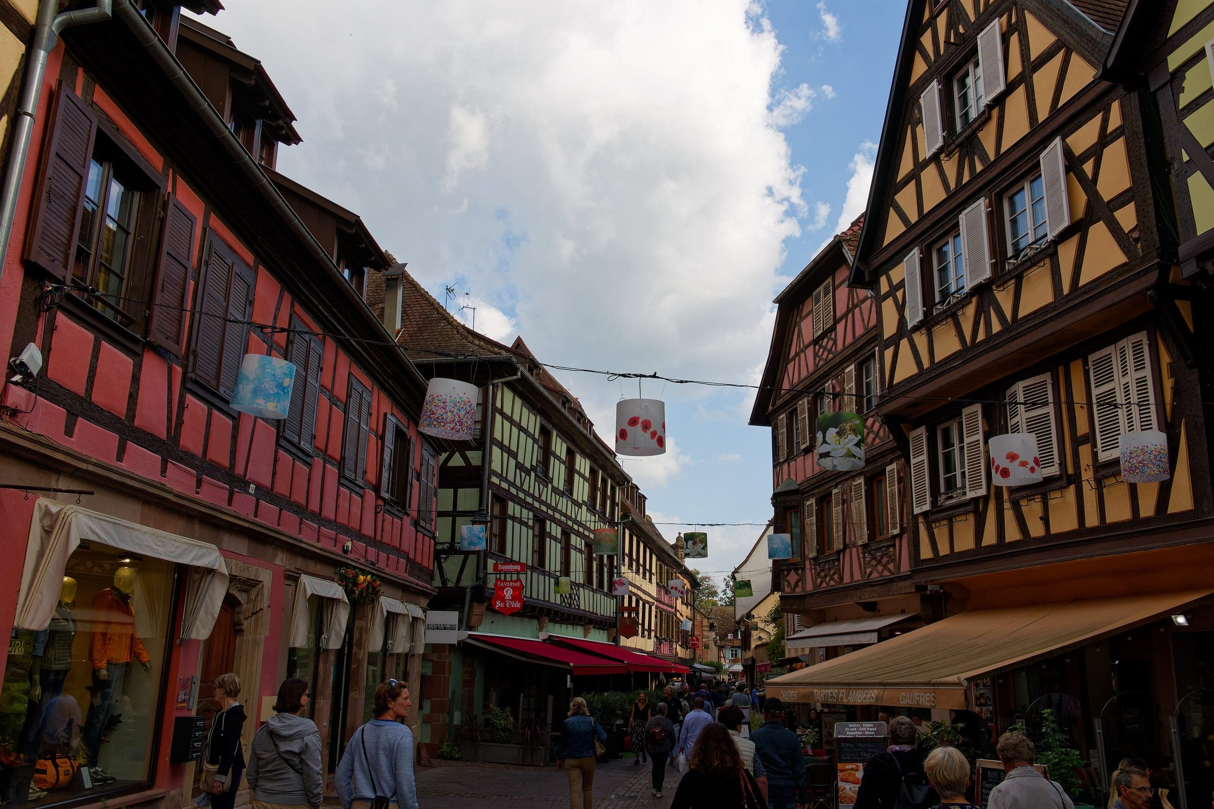 Lanterns hang across a busy shopping street of red and ochre half-timbered houses in Obernai.