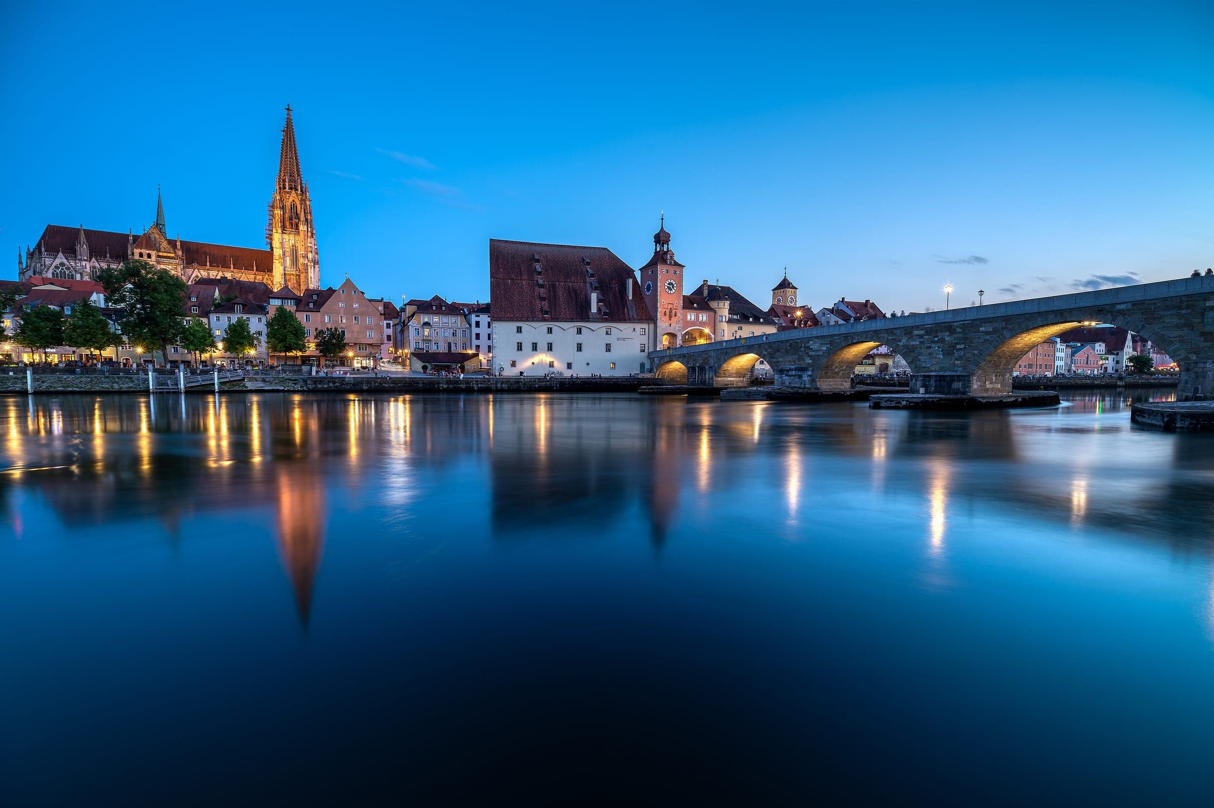 The Stone Bridge and Regensburg Cathedral reflect in the Danube at blue hour with the old town lit.