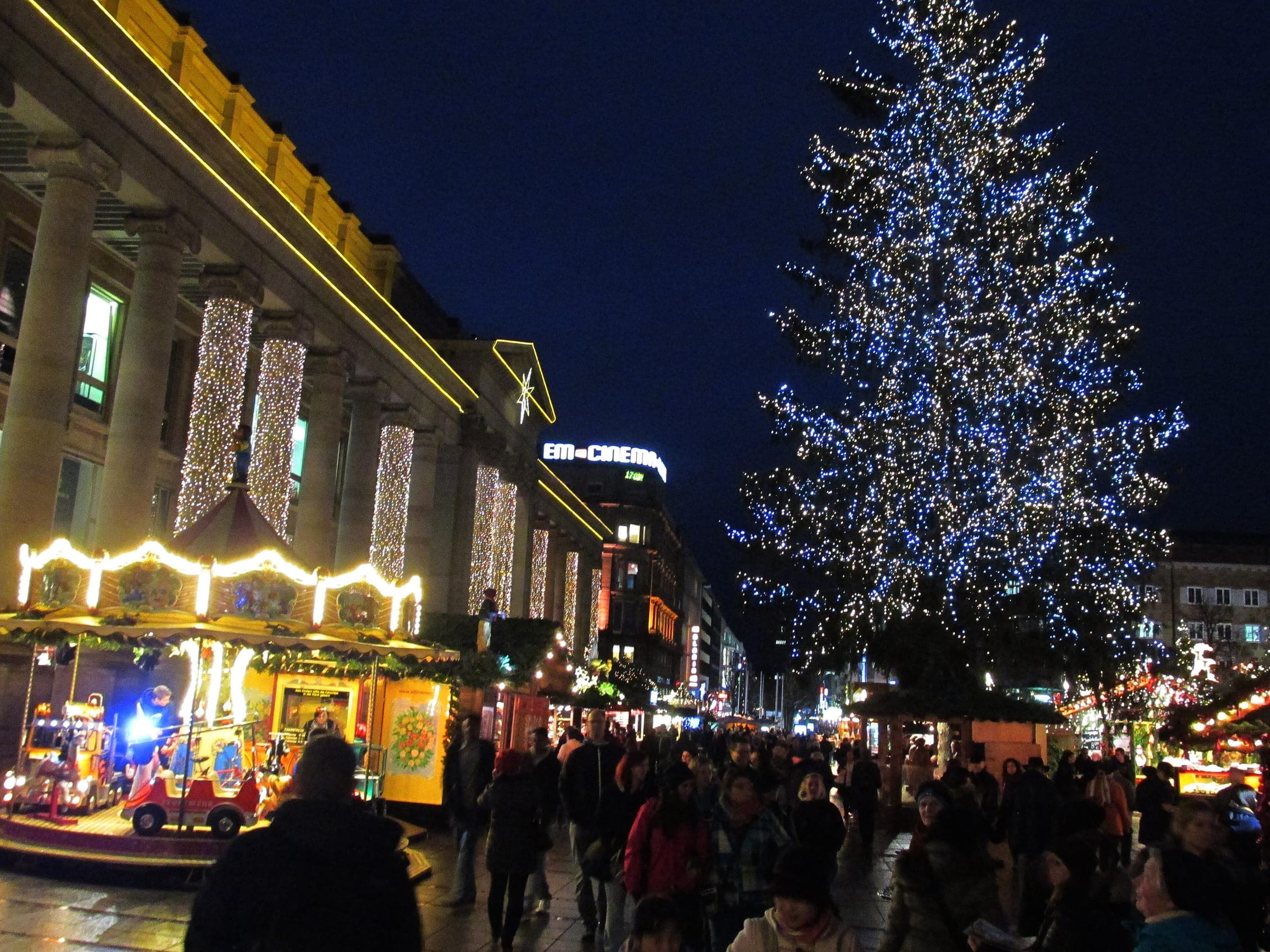 A lit Christmas tree and carousel stand beside the Königsbau colonnade in Stuttgart at blue hour.