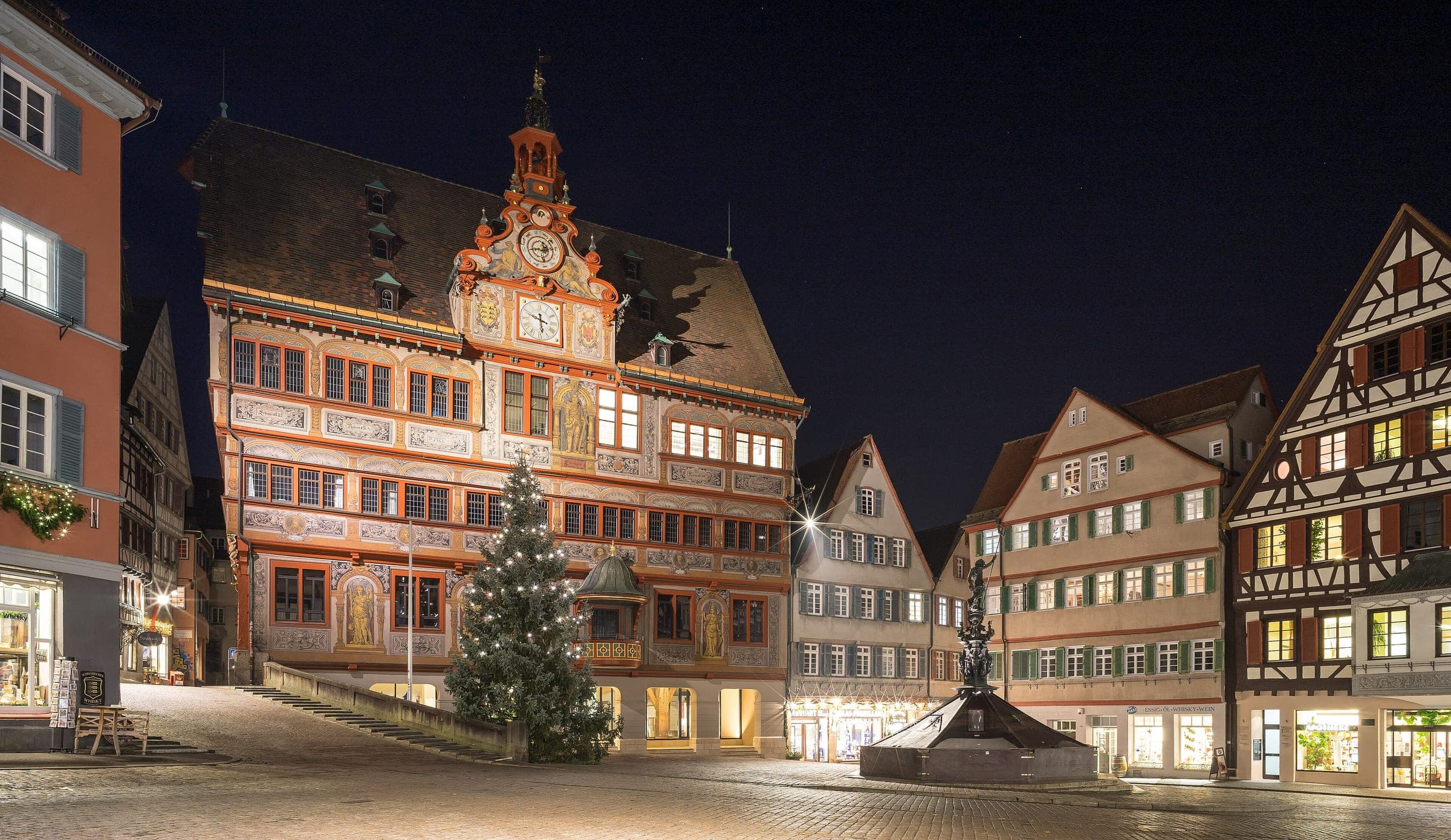 A lit Christmas tree stands before the painted Rathaus on Tübingen's empty Marktplatz on a December night.