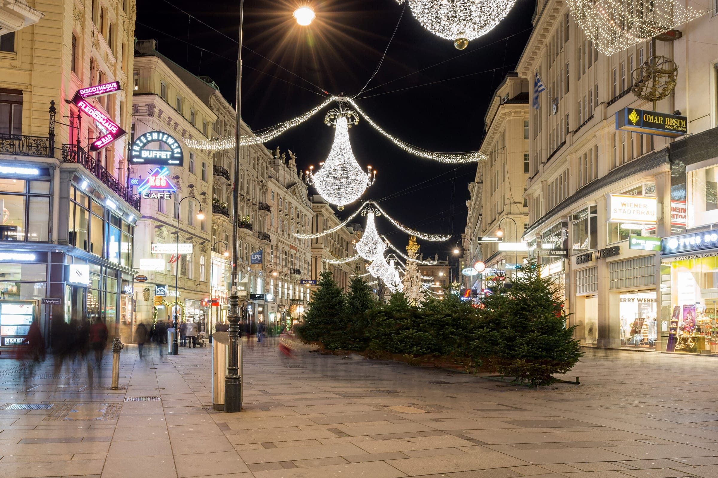 Chandelier lights and a row of small fir trees line a quiet pedestrian street in central Vienna at night.