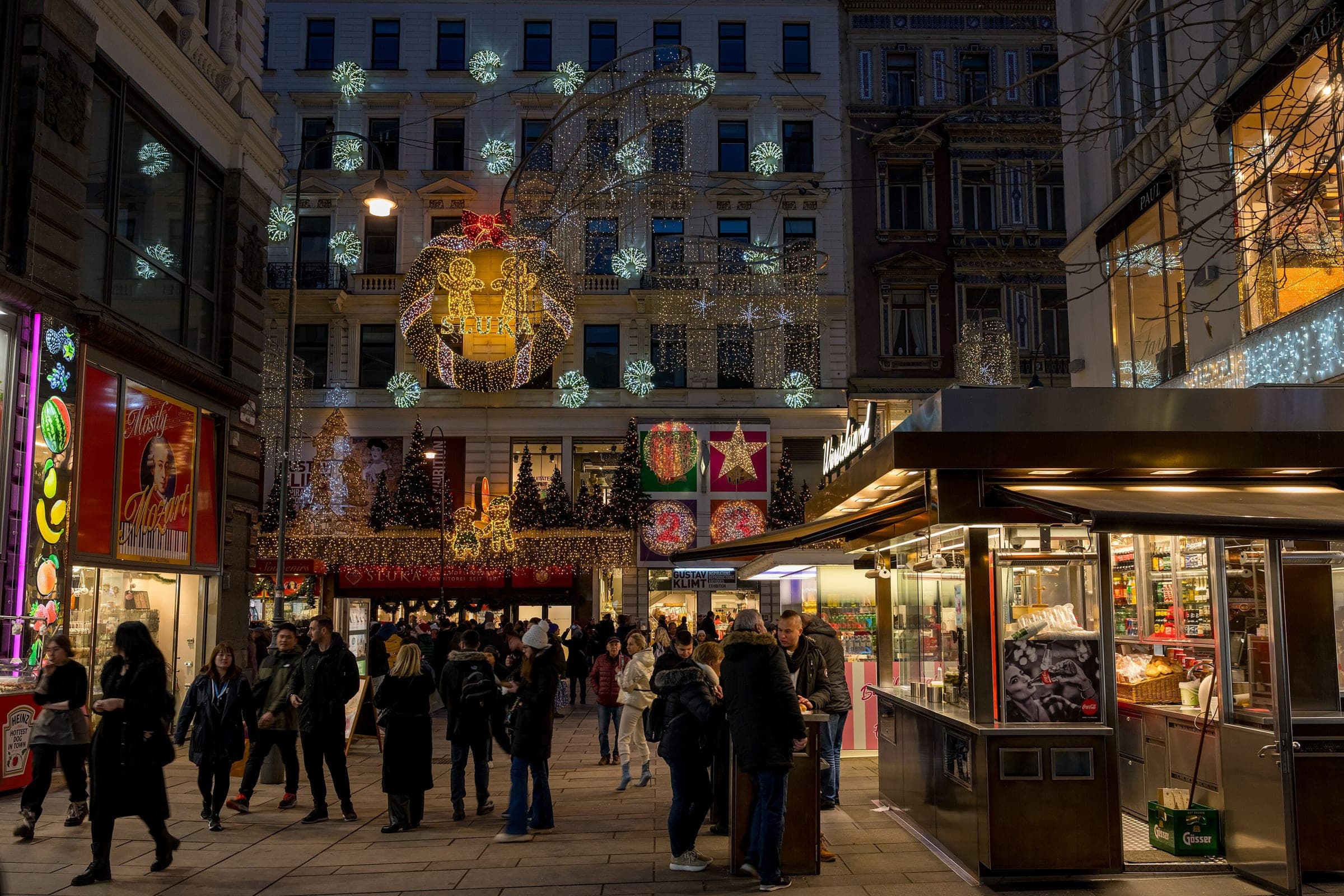 Shoppers walk past a lit sausage stand and glowing star decorations on a street corner in central Vienna.