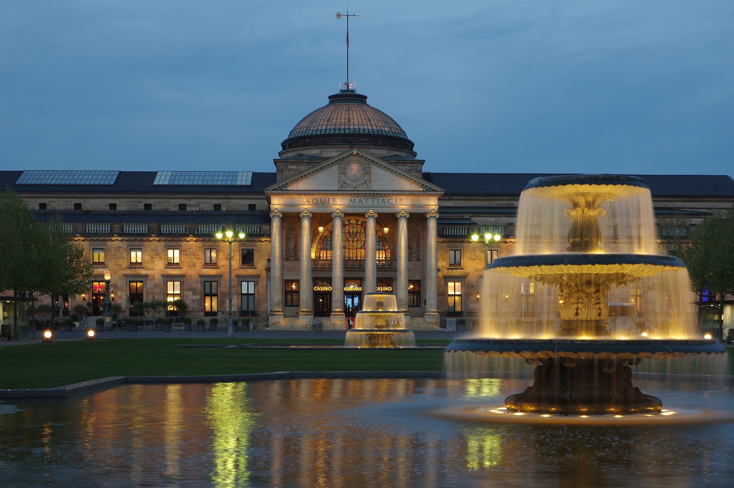 Lit fountains play in front of the Kurhaus in Wiesbaden as blue hour settles over the Bowling Green.