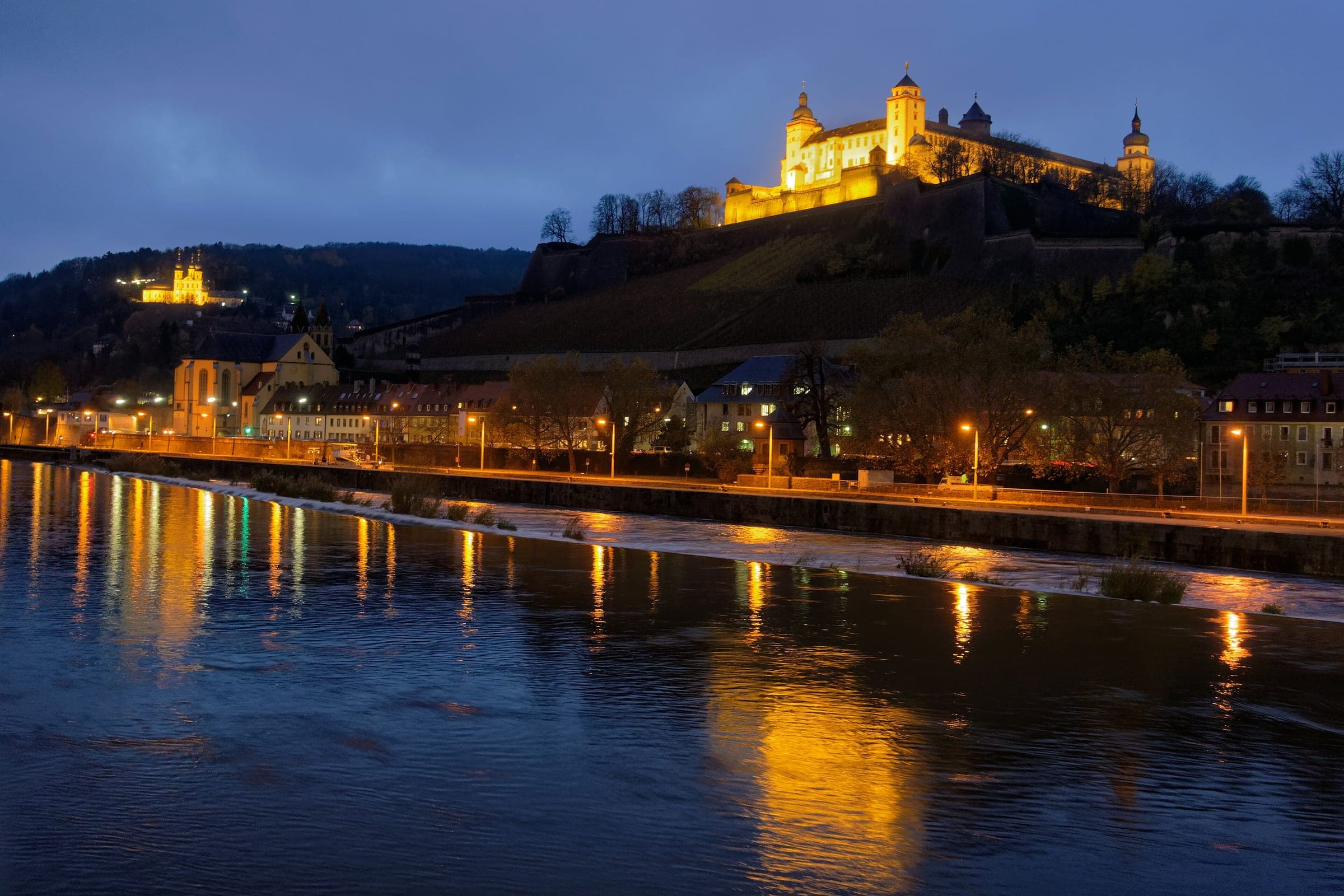 The floodlit Marienberg Fortress shines above the Main in Würzburg at blue hour, with its lights on the river