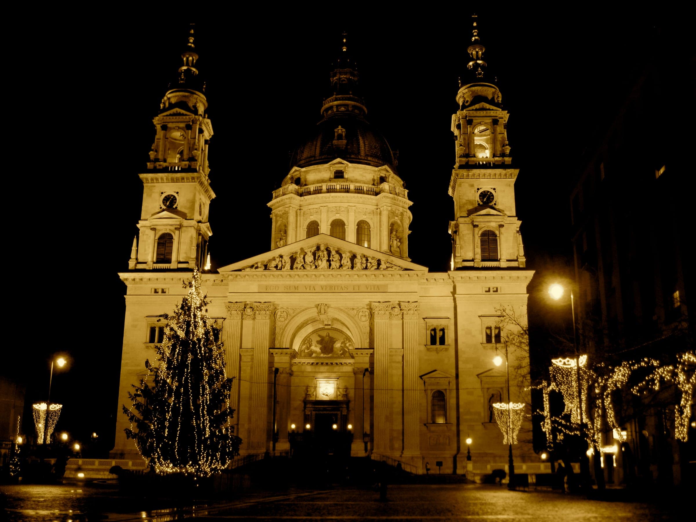 A lit Christmas tree stands before the floodlit facade of St. Stephen's Basilica in Budapest at night.