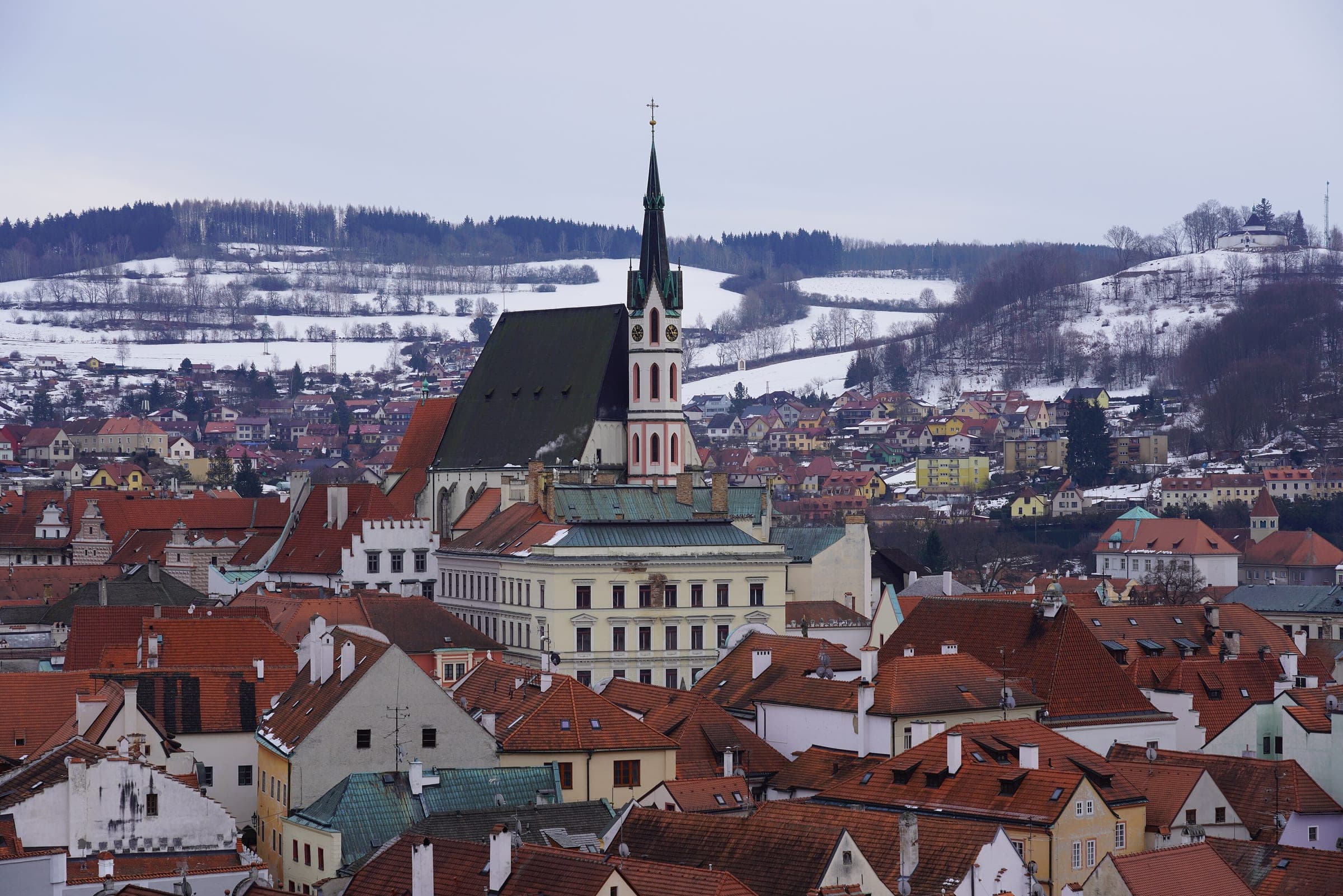 The spire of St Vitus Church rises over the red roofs of Český Krumlov while snow lies on the hills behind.
