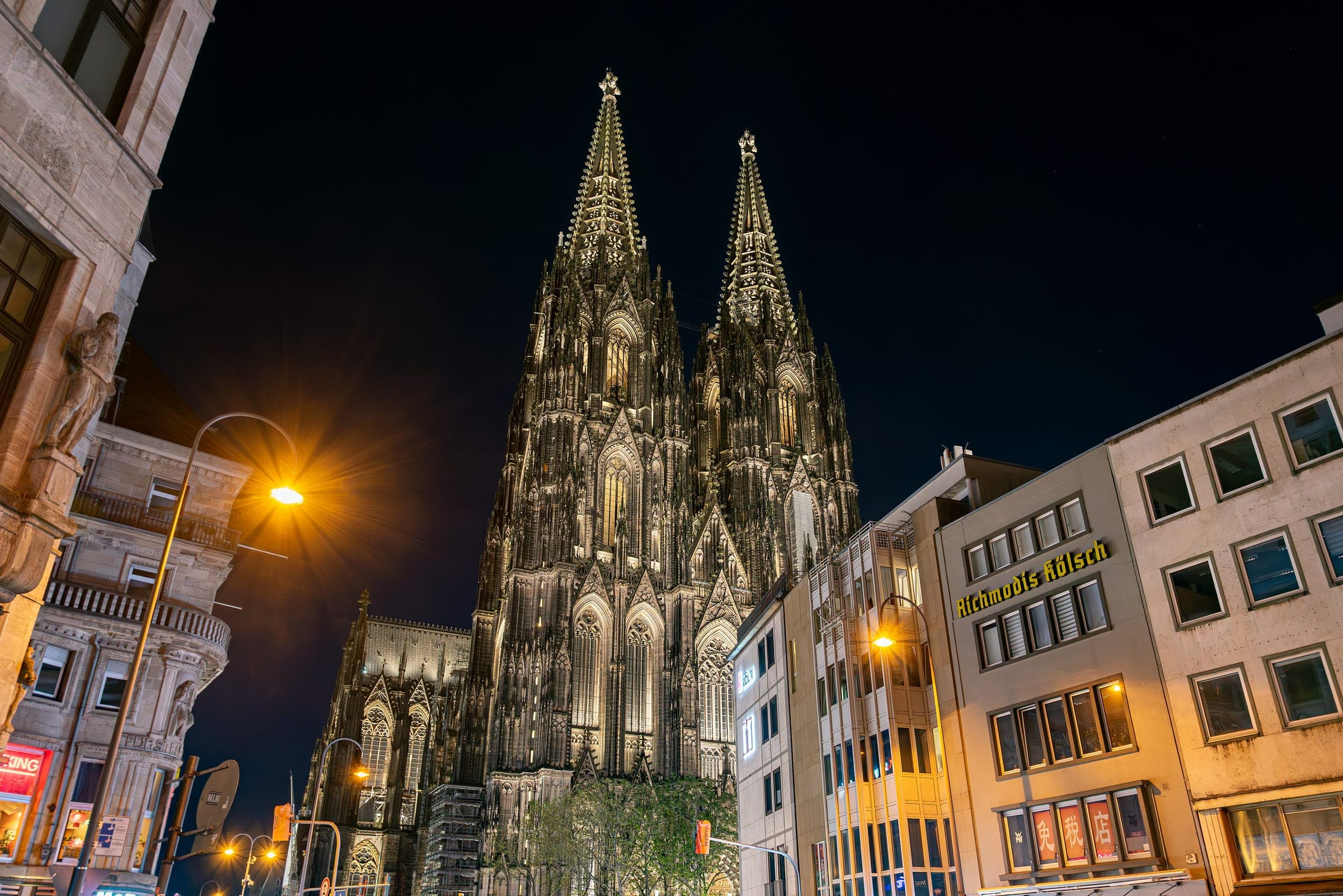 The floodlit west towers of Cologne Cathedral rise over the street at night
