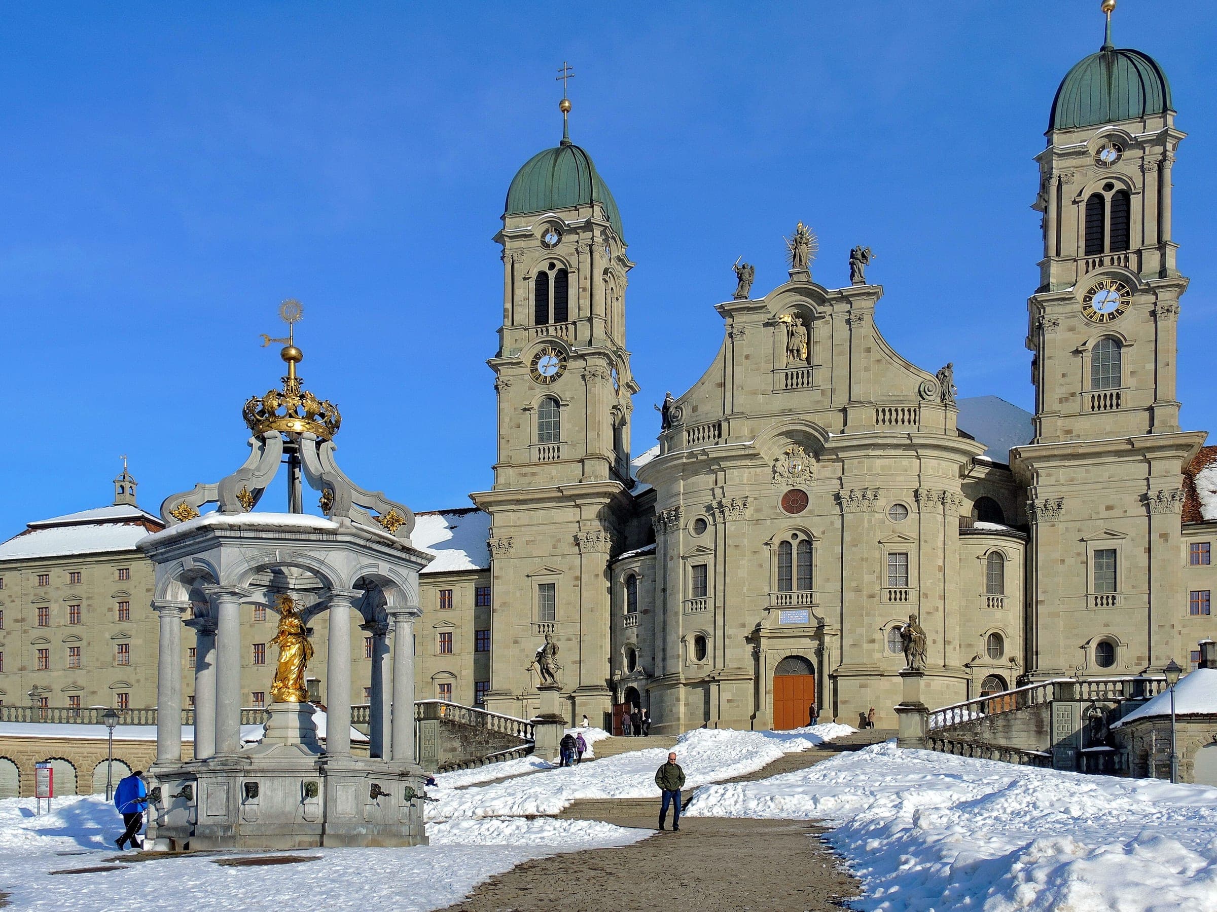 Snow covers the square in front of Einsiedeln Abbey and its Marienbrunnen fountain on a clear winter day