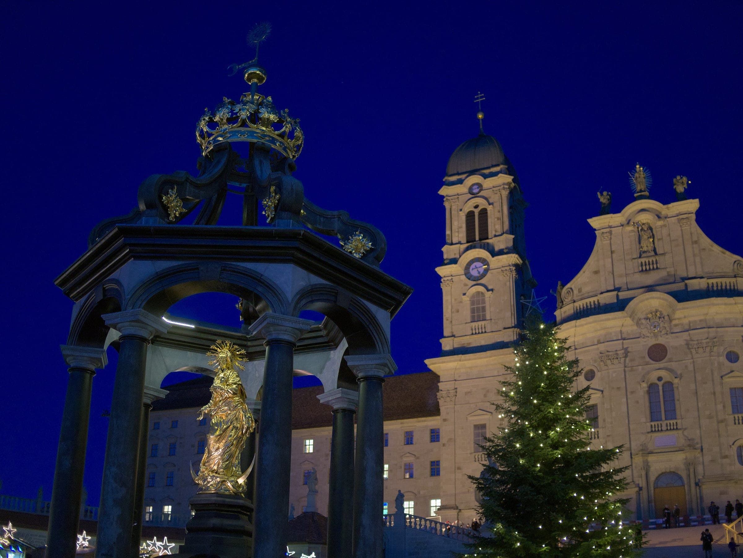 The gilded Marienbrunnen and a lit Christmas tree stand in front of Einsiedeln Abbey at blue hour