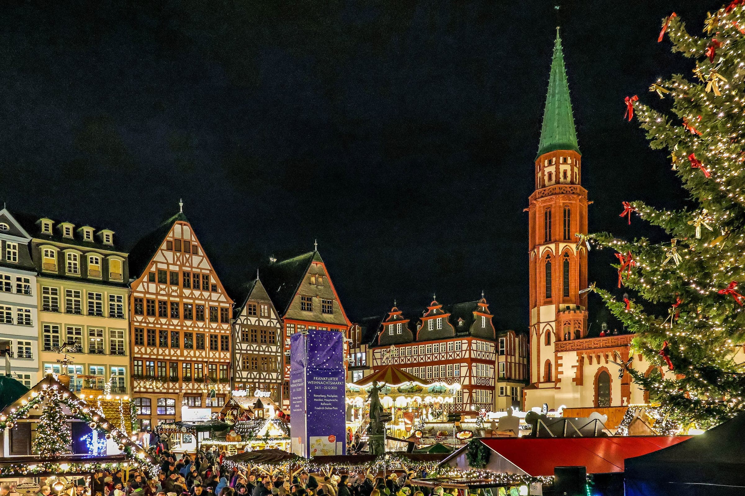The red tower of the Alte Nikolaikirche rises over the Ostzeile houses and market stalls on the Römerberg at night.