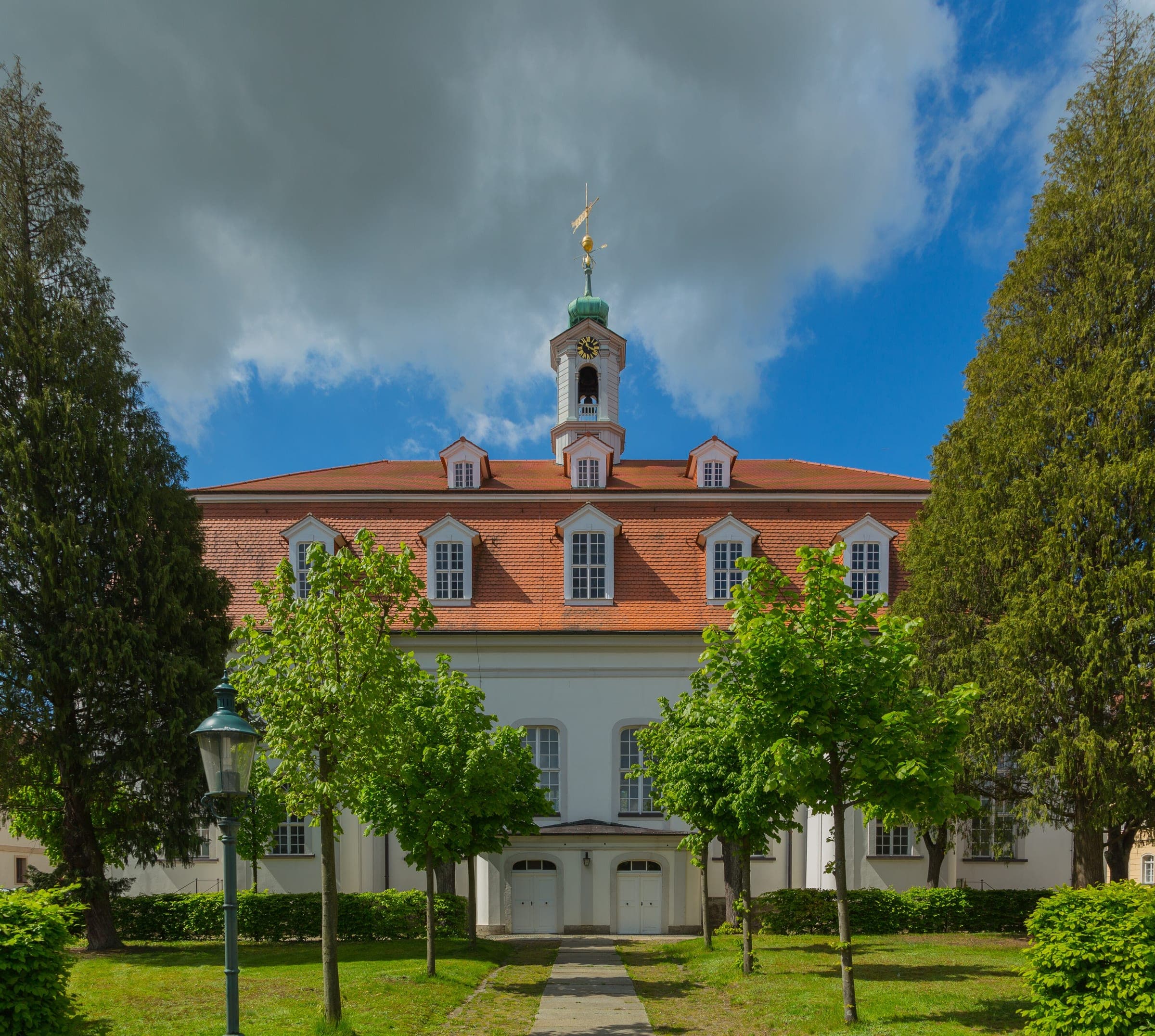 A tree-lined garden path leads to the white facade and small bell tower of the Kirchensaal in Herrnhut.