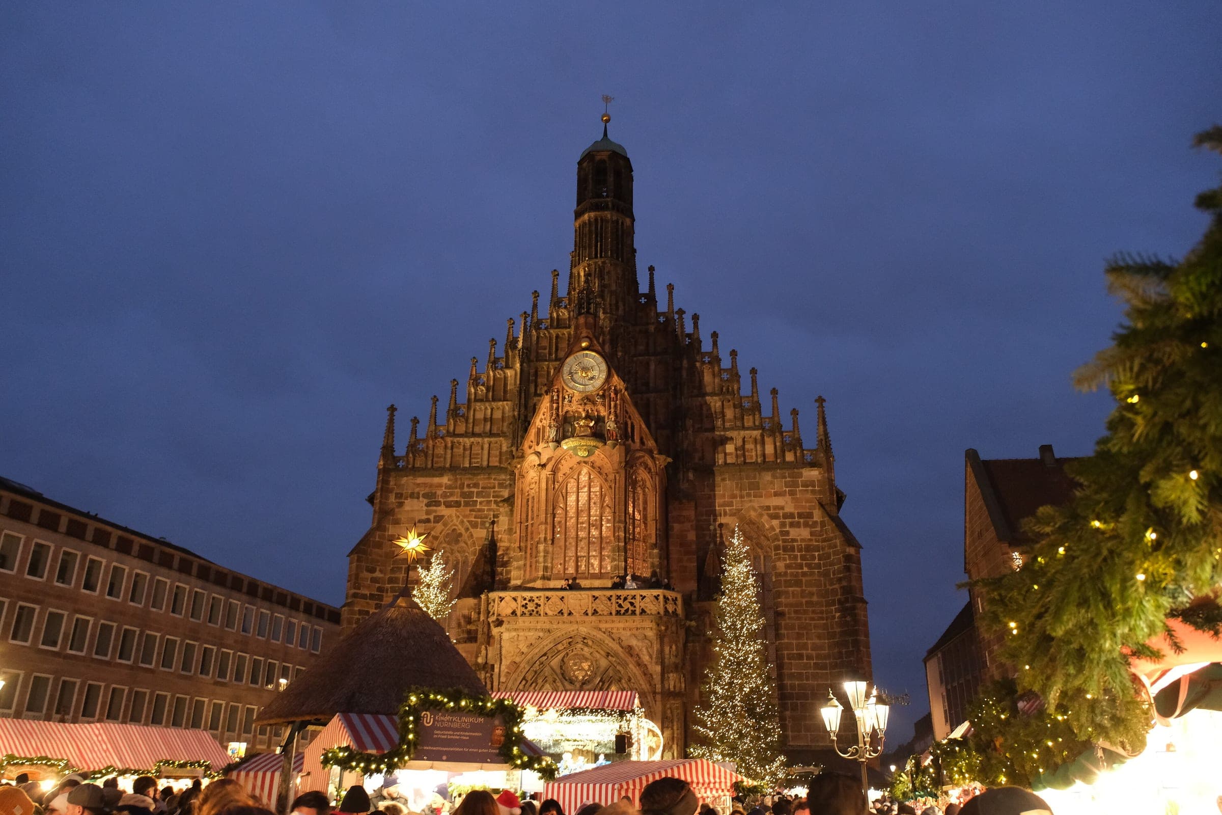 The Frauenkirche stands over the Hauptmarkt at blue hour, with a lit tree and market stalls at its feet