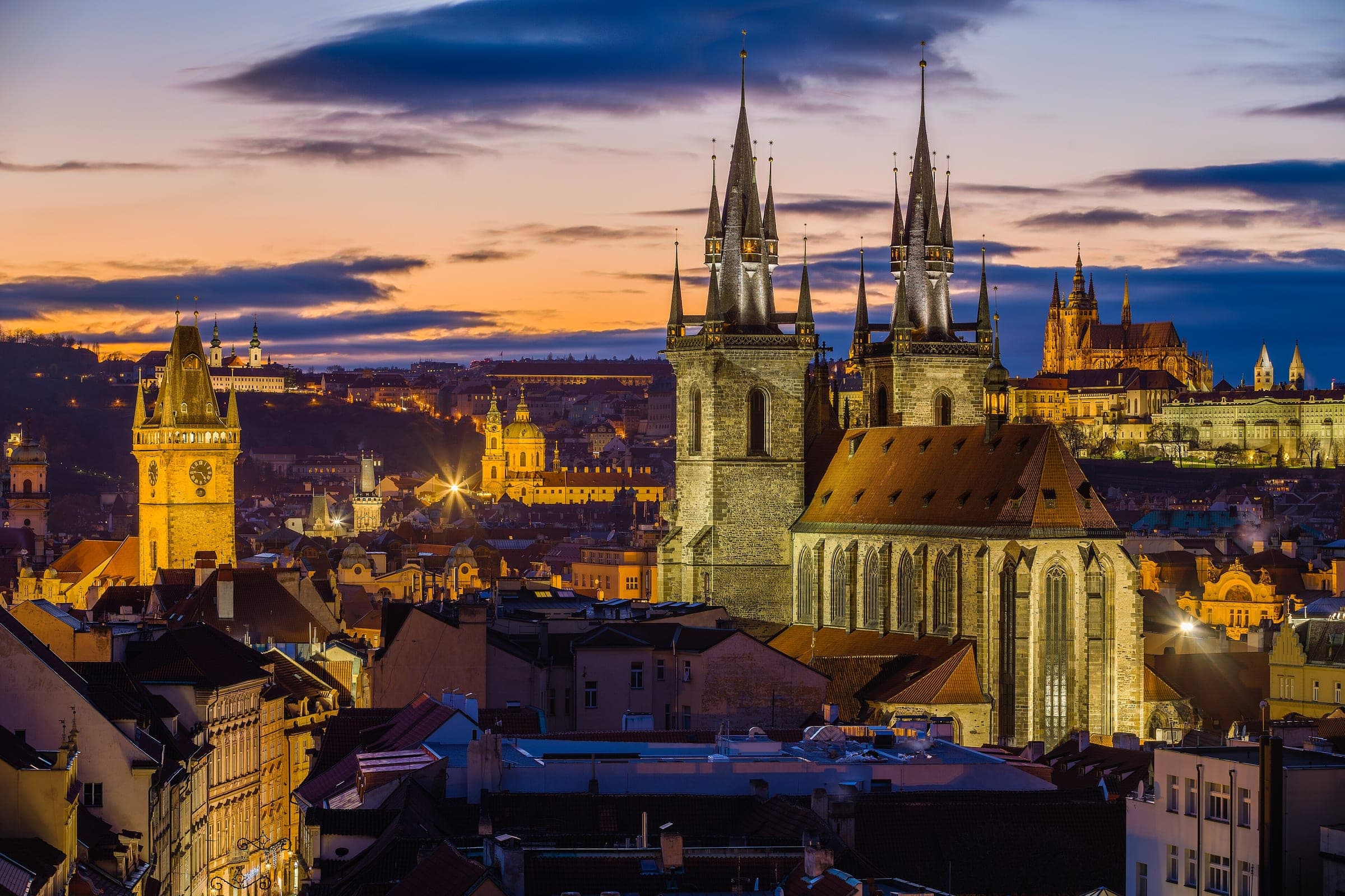 The Týn Church spires rise over the Old Town roofs at dusk while Prague Castle glows on the far hill.