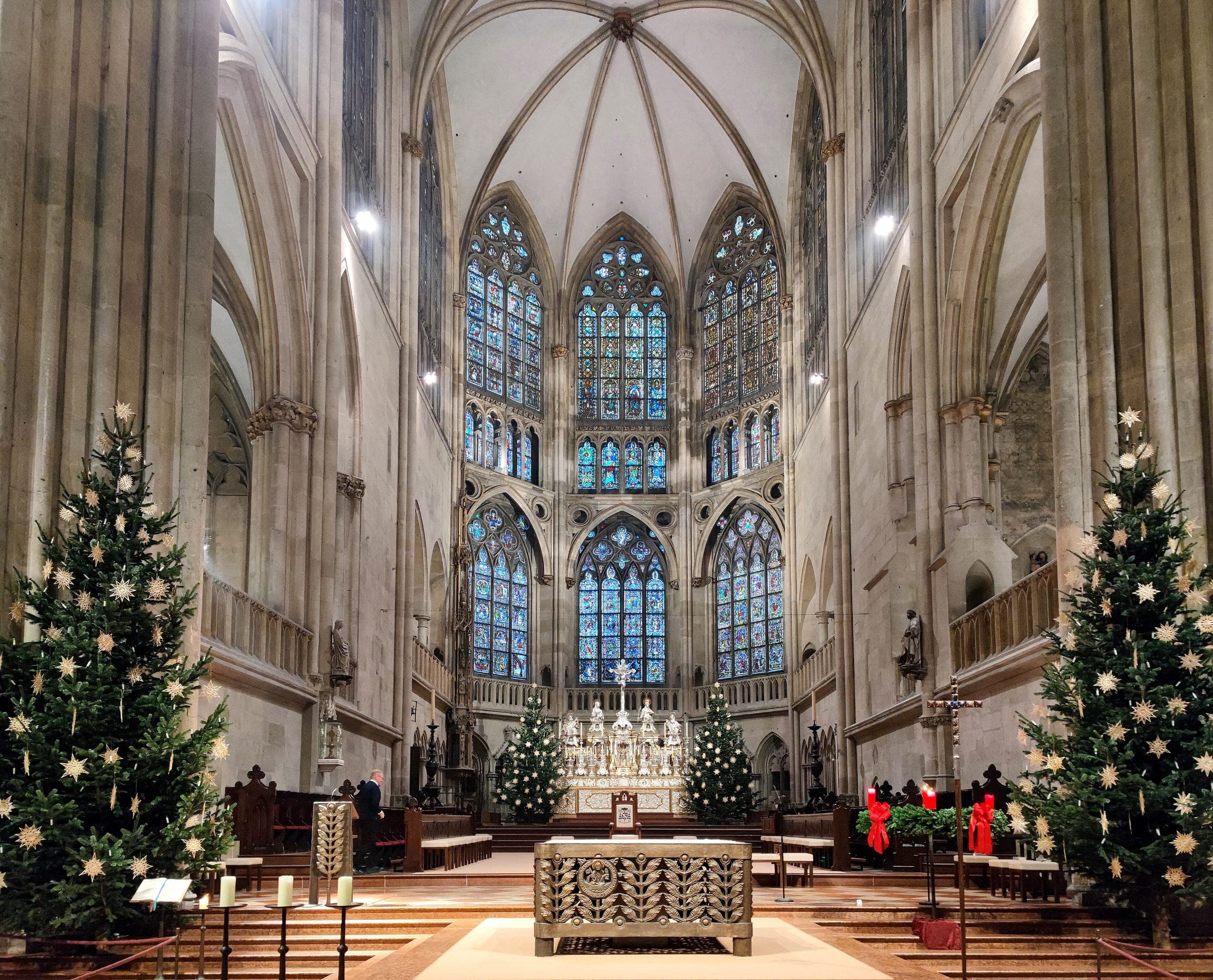 Christmas trees with straw stars flank the chancel of Regensburg Cathedral under its tall stained glass windows.
