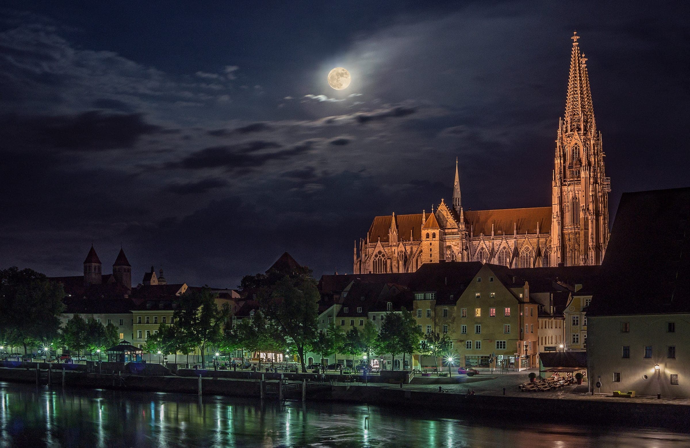 A full moon rises over the floodlit spires of Regensburg Cathedral above the Danube at night.