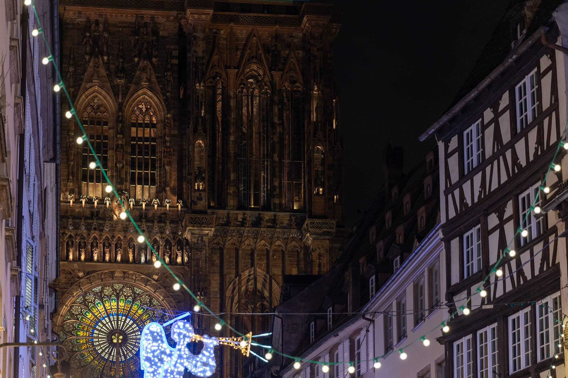 Strasbourg Cathedral's rose window glows at night above a lit angel and garlands strung across rue Mercière.