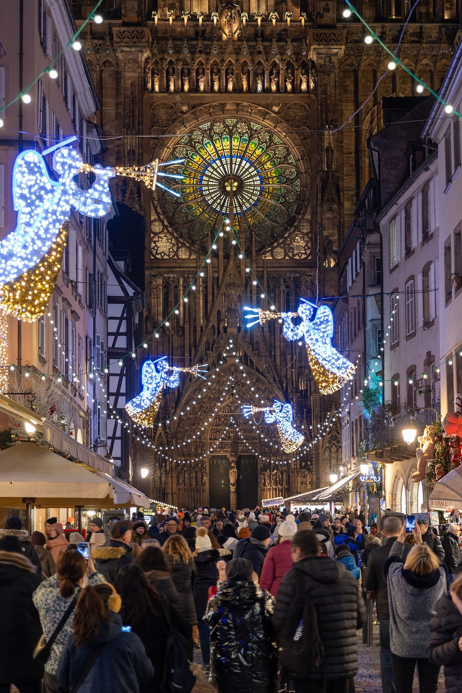 Crowds walk up rue Mercière under lit angels toward the west front of Strasbourg Cathedral at night.
