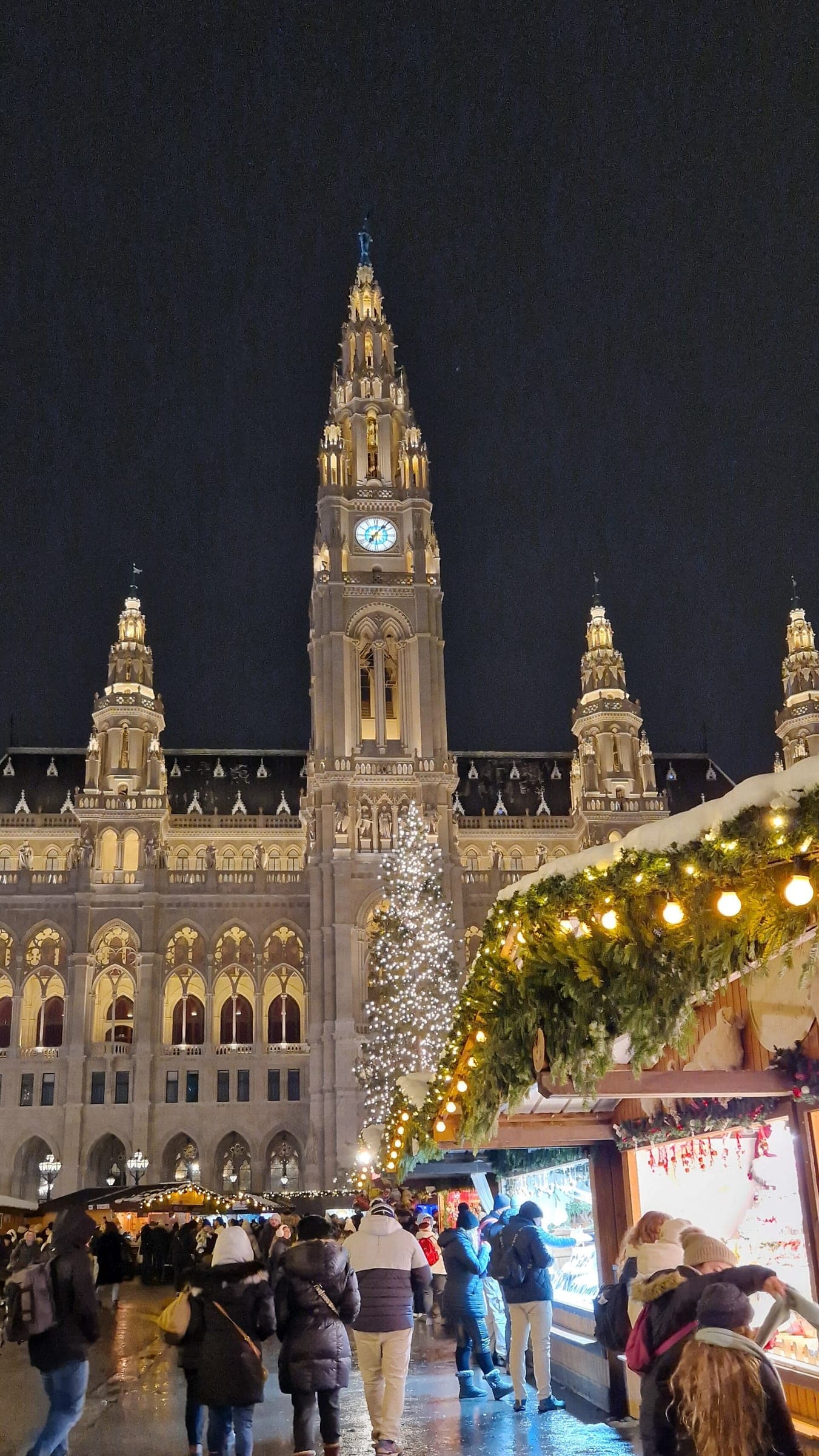 The clock tower of Vienna City Hall rises above a lit tree and snowy market stall roofs on a winter night.